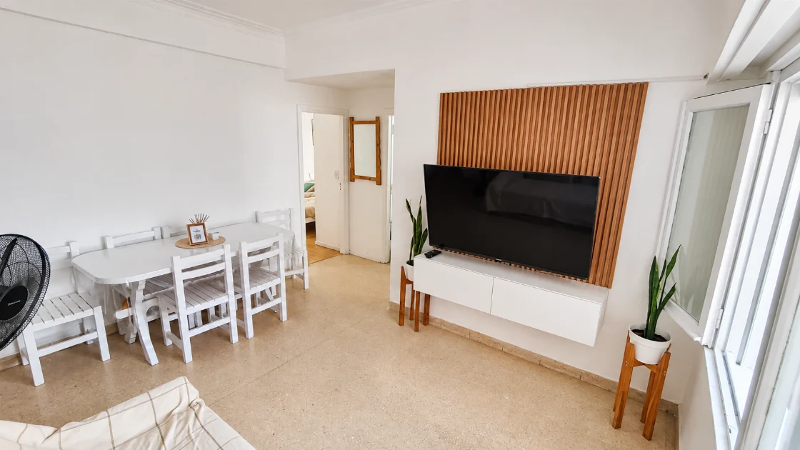 Living-comedor con mesa y sillas blancas, mueble de TV y ventana.